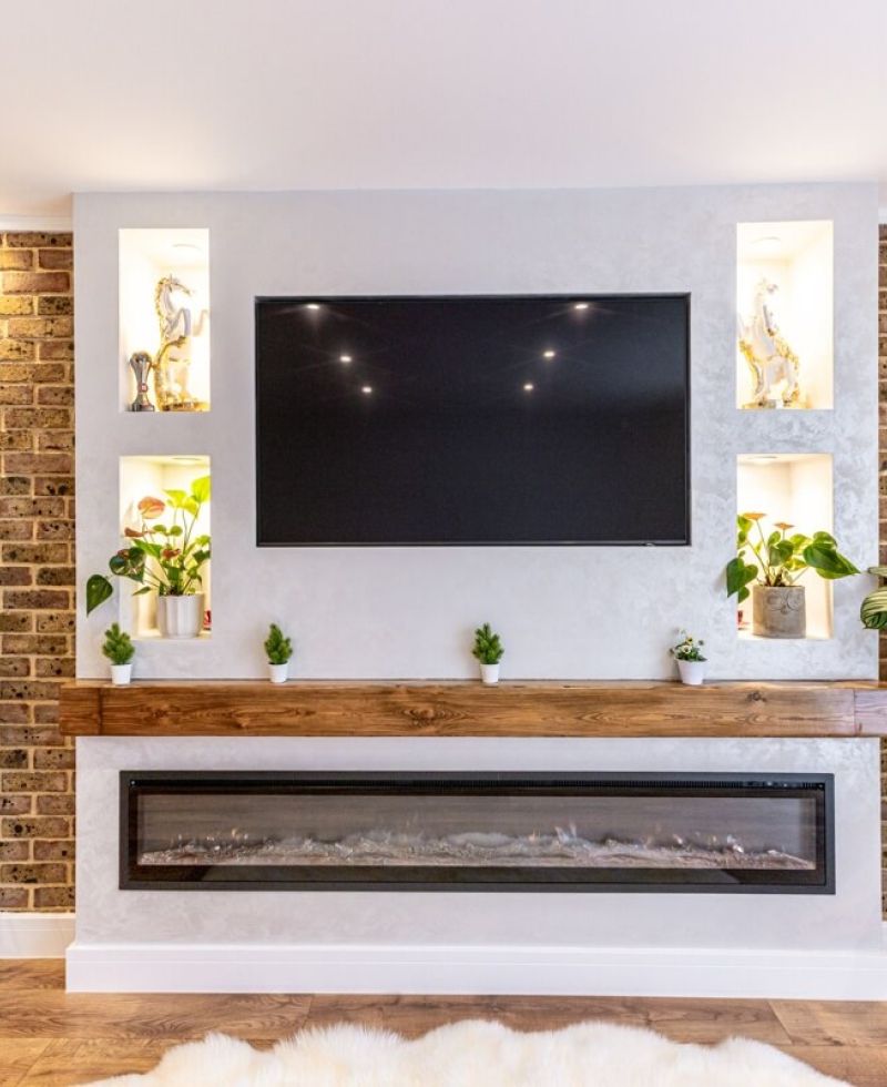 Modern living room feature wall with a large flat-screen TV mounted above a sleek linear electric fireplace, framed by exposed brick accents and built-in illuminated display niches.